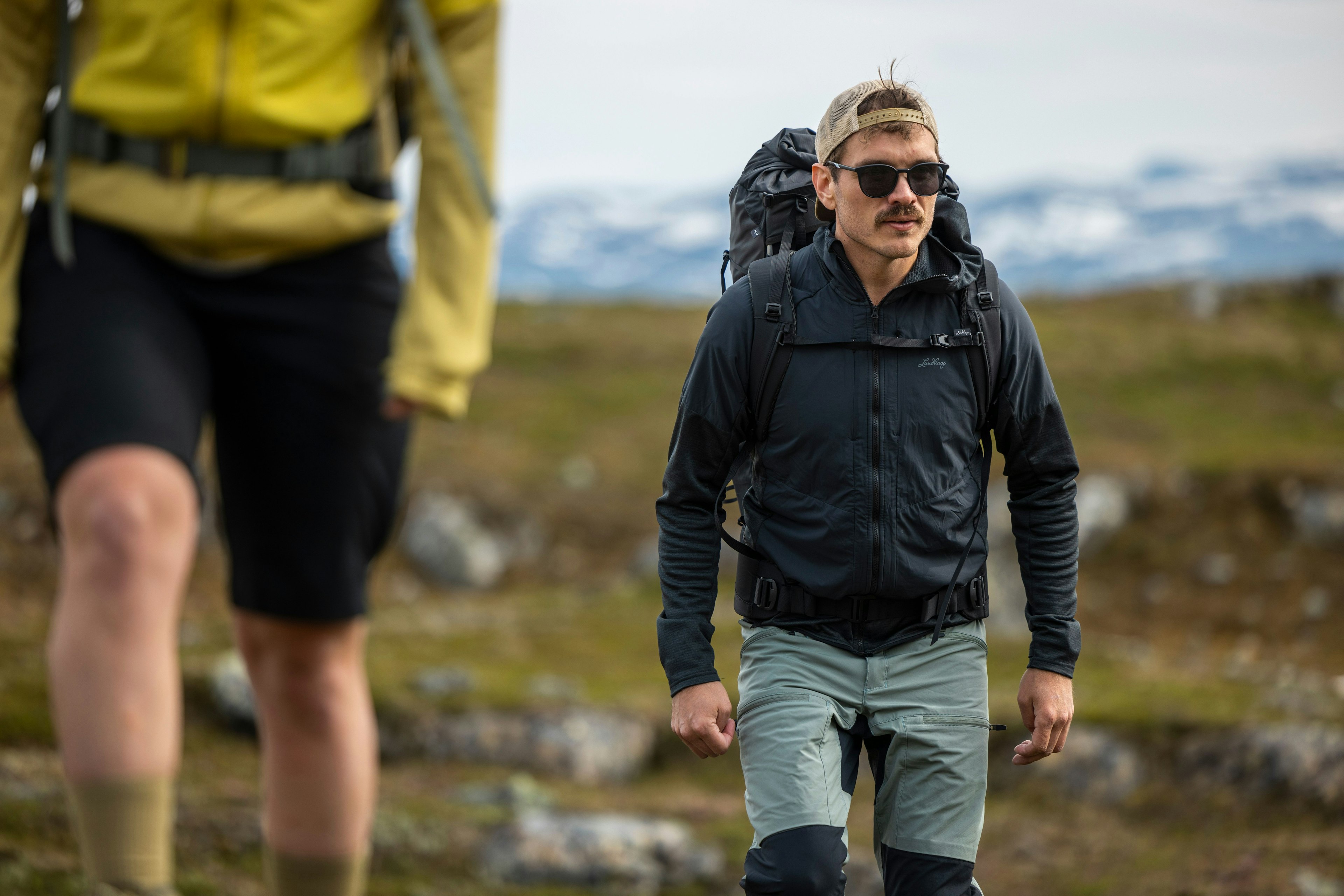 To personer på fottur i et fjellandskap, med en mann i forgrunnen iført en svart jakke, grå bukser og en ryggsekk, mens en annen person i gult er uskarp i bakgrunnen.