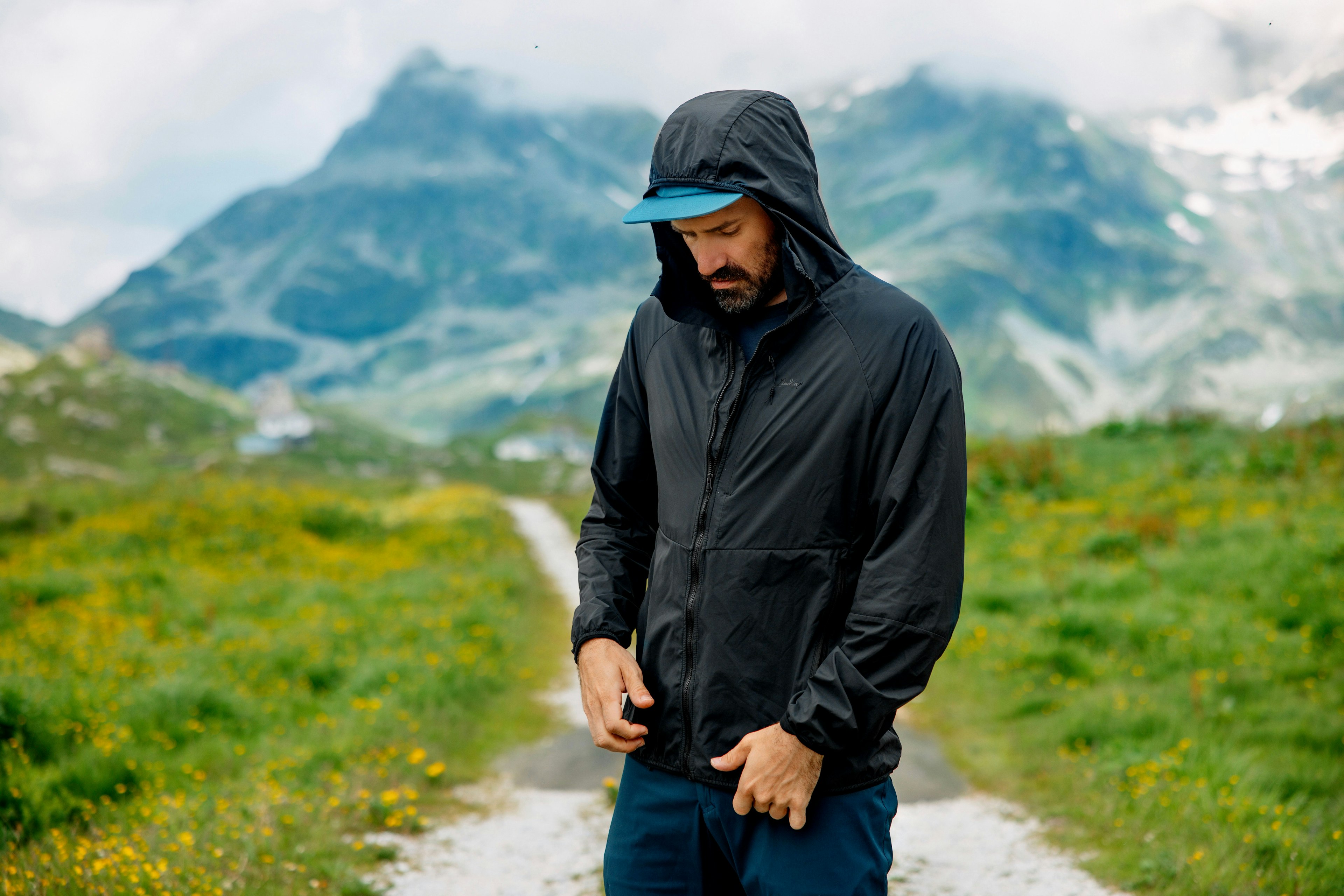 A man in a black shell jacket and blue cap stands on a mountain trail, adjusting his jacket in a natural alpine setting.