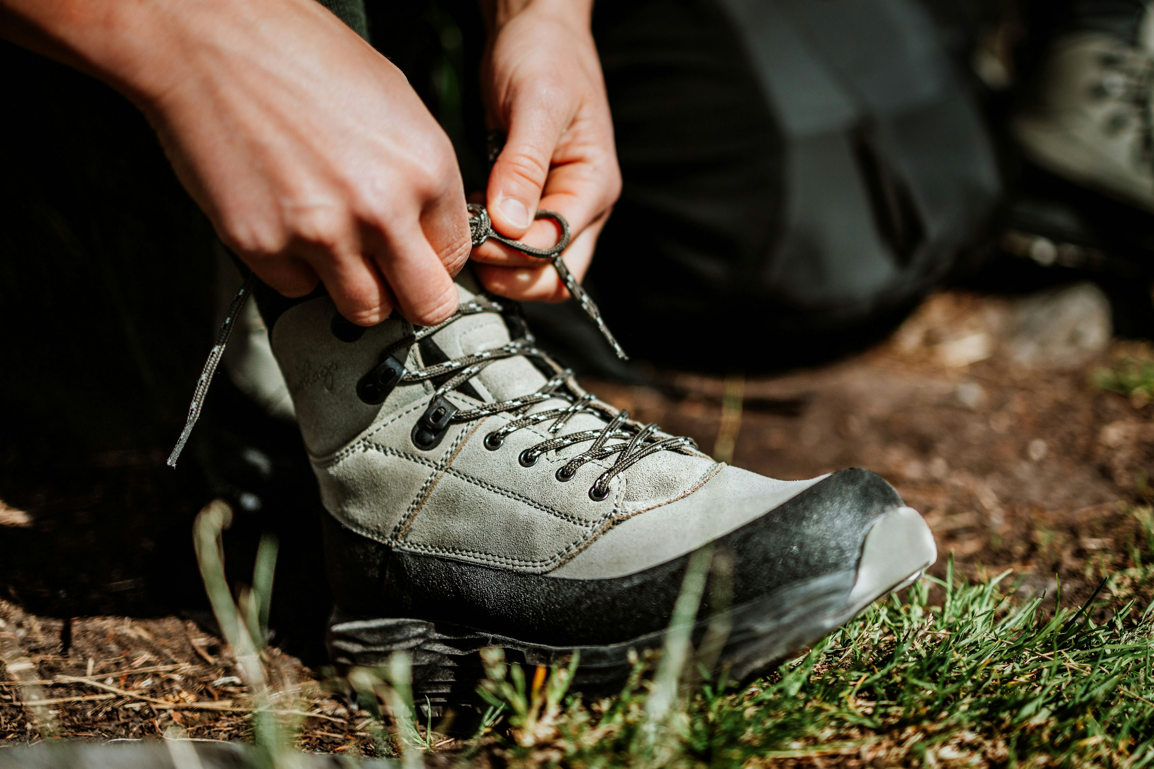 Nærbilde av hender som snører en Lundhags Tived Trail Boot på naturlig underlag utendørs.