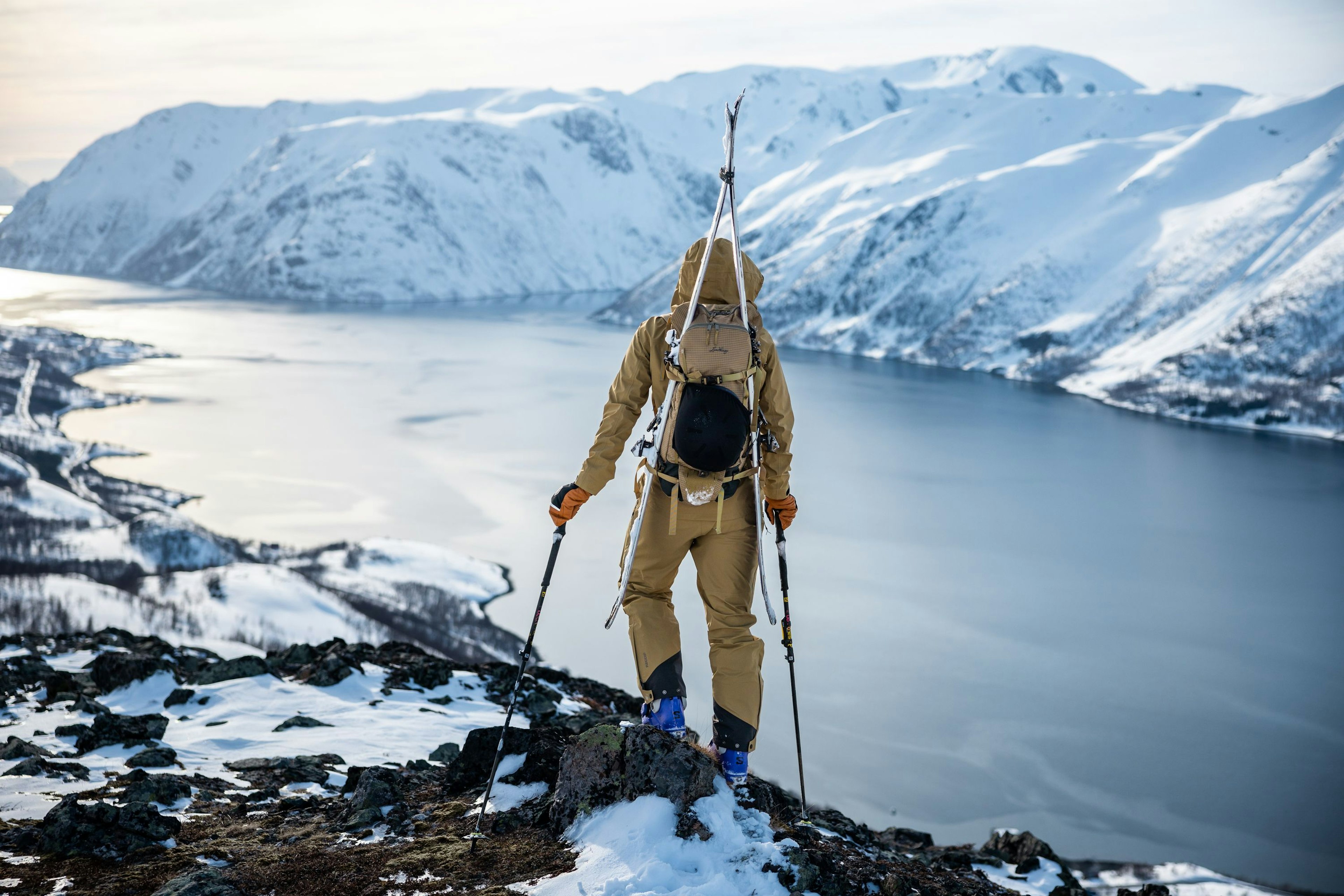 Person klädd i Lundhags Abisku Waterproof för topptur med skidor på ryggen, stående på en klippig, snötäckt bergsrygg med utsikt över en fjord och snöklädda berg.