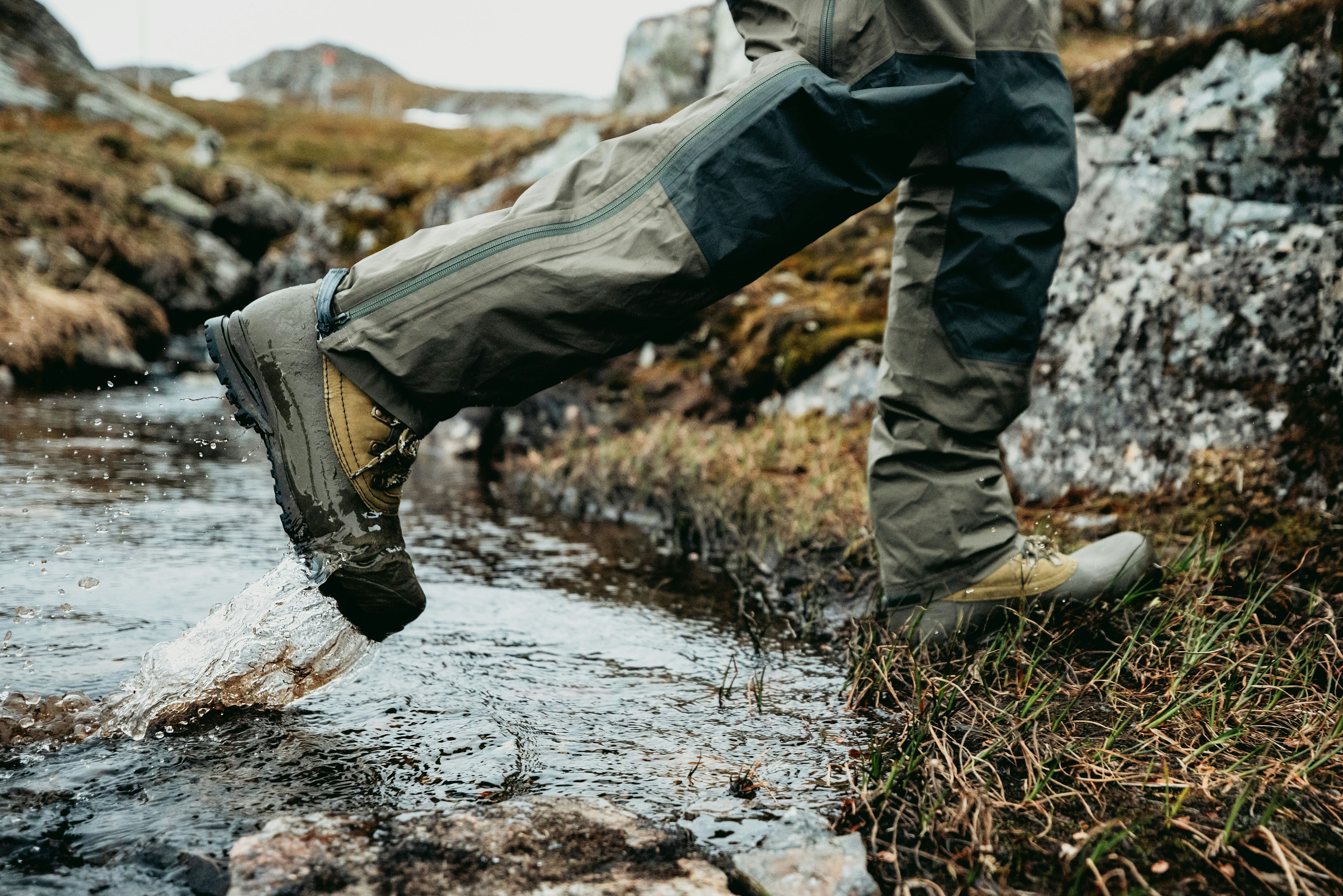 Närbild av en person som krysser en grunn bekk i krevende terreng, iført vanntette tursko og forsterkede skallbukser.