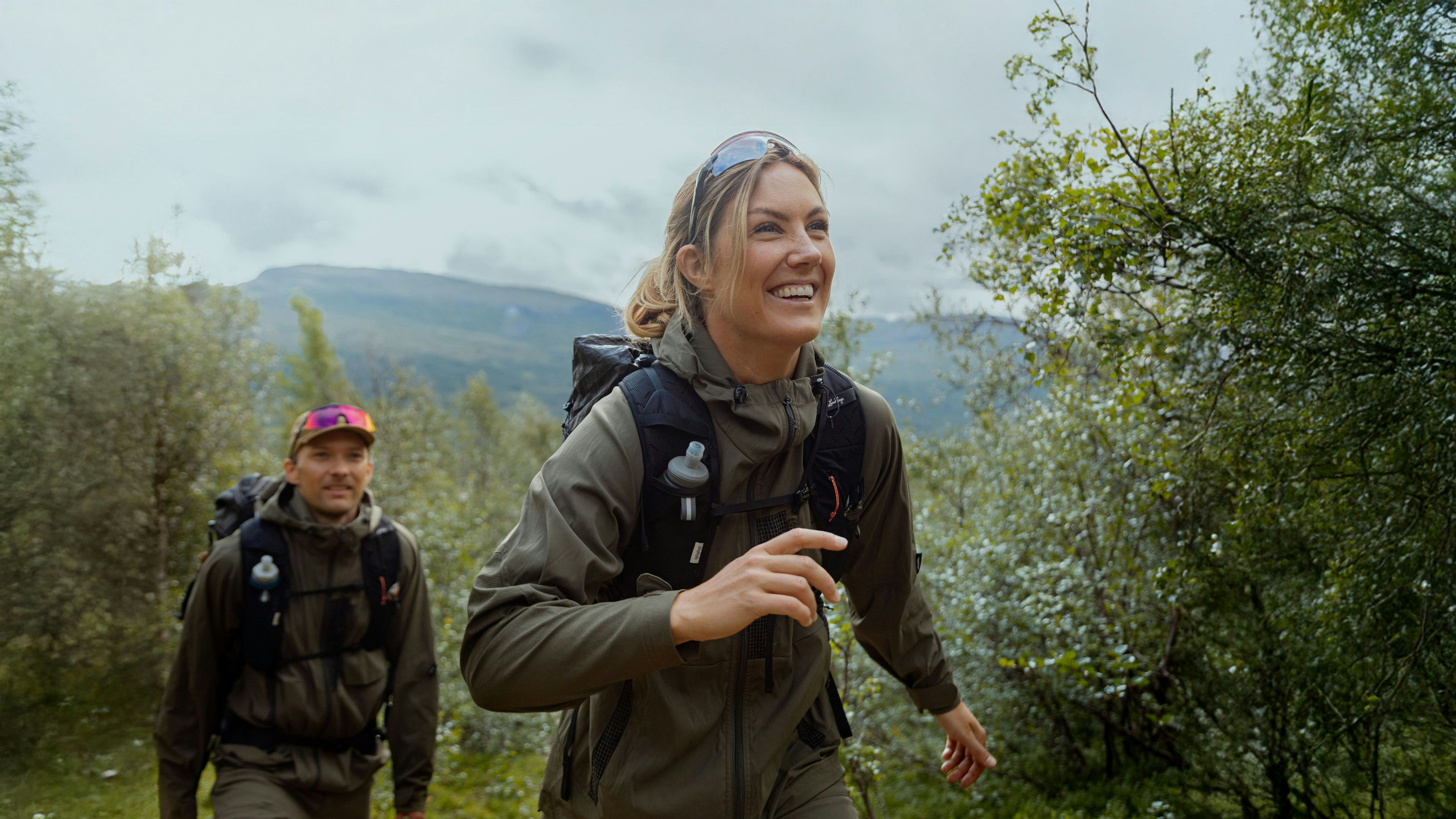 Three people ski touring uphill on a snowy slope with dramatic mountain peaks and a fjord in the background, under a partly cloudy sky.