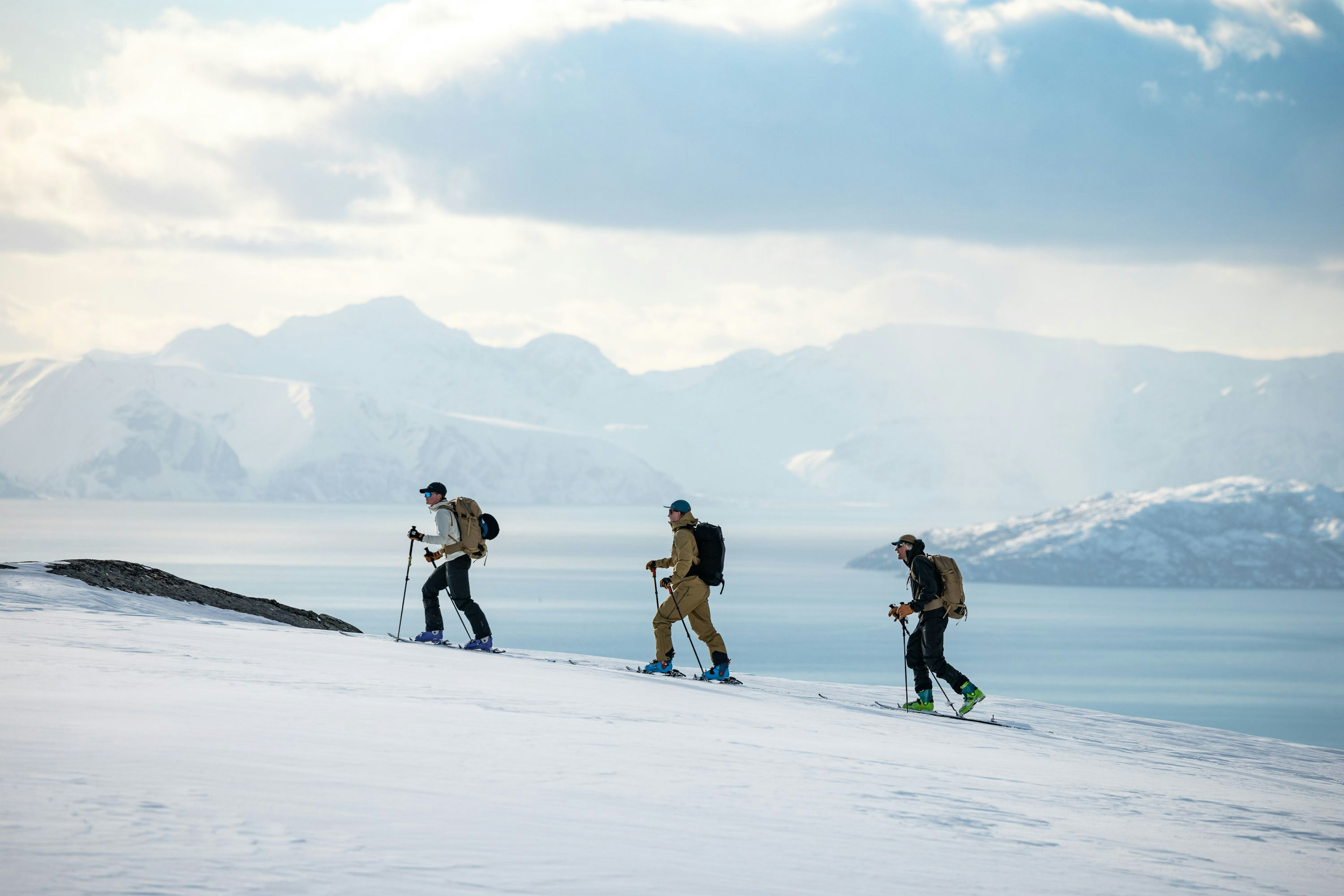 Three people ski touring uphill on a snowy slope with dramatic mountain peaks and a fjord in the background, under a partly cloudy sky.